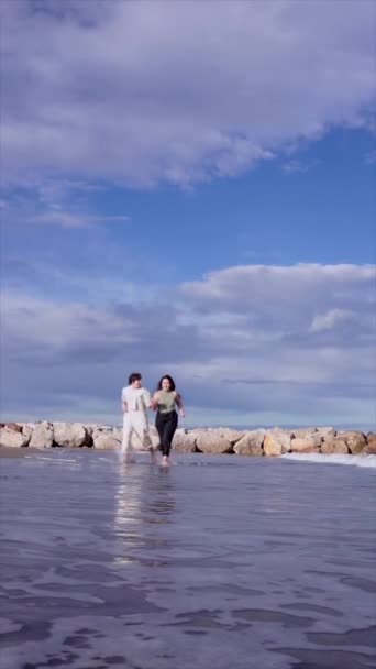 笑顔で陽気な同性愛女性の下からカジュアルな服を着て 晴れた日に海の水を飛ばして互いを見つめ合う手を握る — ストック動画