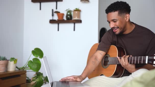 ポジティブなアフリカ系アメリカ人の男性ミュージシャンは グレーの背景に対して快適なソファーでアコースティックギターを再生しながらラップトップ上のビデオチュートリアルを見ています — ストック動画