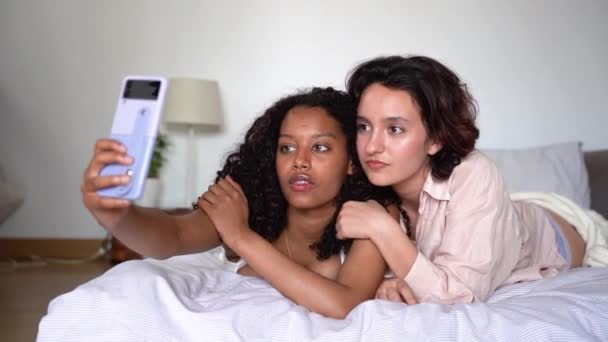 若いアフリカ系アメリカ人女性と笑顔の多様な友人が自宅でホワイトベッドに横たわり 週末を楽しんでいる間に携帯電話で自己肖像画を撮る — ストック動画