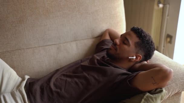 非洲裔美国男子躺在舒适的沙发上 闭上了眼睛 一边在家里用真正的无线耳塞听音乐 一边松口气 — 图库视频影像
