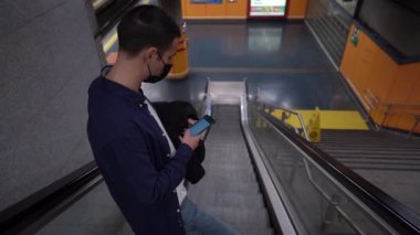 Yukarıdan, maskeli, metroda merdivenlerde yürürken cep telefonunun ekranına dokunan kimliği belirsiz genç bir adam görünüyor.