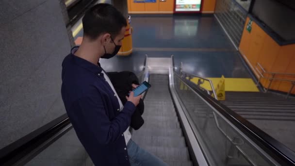 地下鉄の階段に立ちながら携帯電話の画面に触れる顔マスクの匿名の若い男性の側のビューの上から — ストック動画