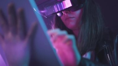 Modern VR kulaklık takan konsantre olmuş kadın karanlık odada duvarın yanında duruyor ve çağdaş tablet neon ışığı altında.