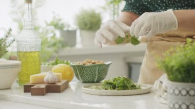 Steril eldiven ve önlüklü tanınmayan bir kadın taze fesleğen yapraklarını tabağa koyarken gün ışığında mutfakta sağlıklı bir akşam yemeği hazırlıyor.