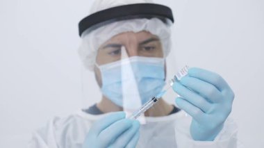 Ekin, modern donanımlı hastanede çalışırken cerrah önlüğü eldivenleri ve yüz kalkanı ile şırıngayı şişeden aldığı ilaçlarla dolduran profesyonel erkek tıp doktoru.