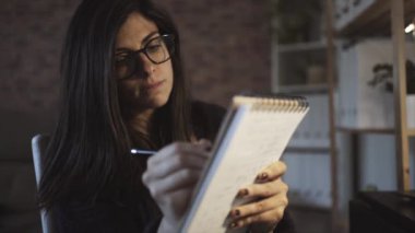 Gözlüklü konsantre kadın bestecinin uzaktan çalışma sırasında dizüstü bilgisayarla masada otururken not alırken yan görünümü