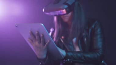 Modern VR kulaklık takan konsantre olmuş kadın karanlık odada duvarın yanında duruyor ve çağdaş tablet neon ışığı altında.
