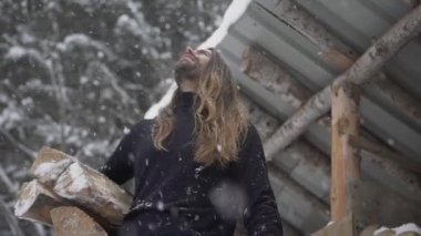 Uzun saçlı, sıcak kazak giyen, elinde odun tutan yakışıklı bir erkek. Kışın barakanın yanında dururken kar tanelerinin düşüşüne huzur içinde bakıyor.