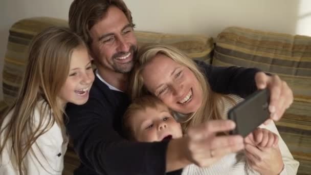 笑顔の妻と可愛い子供たちと一緒にソファに座って携帯電話で自撮りしながら抱きしめる陽気な大人の男の高角度 — ストック動画
