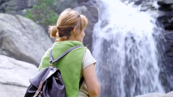 成熟した女性ハイカーでカジュアル服でバックパックを着て森の中に立っている強力な滝の景色を賞賛 — ストック動画