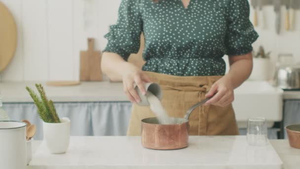 キッチンのテーブルで食べ物を準備しながら ポットにセモリナ粉のカップを追加する匿名の女性料理を作る — ストック動画