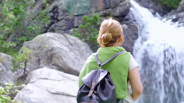 バックパック付きのカジュアルな服で匿名の女性ハイカーのビュー強力な滝の景色を賞賛森の中に立って — ストック動画