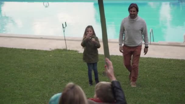 幸せな大人親とともに小さな息子と娘で暖かい服遊びますボールとともに草の芝生の上でプールサイド — ストック動画