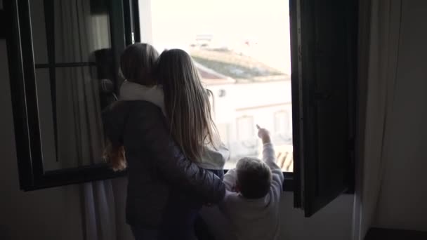 可愛い息子と一緒に窓の外を眺めながら笑顔と小さな娘を抱きしめる暖かい服で陽気な若い女性 — ストック動画