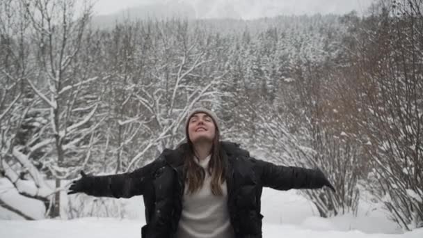 暖かい服と興奮して腕を伸ばし 雪の中で凍った森の中で寒い冬の日を過ごしている間に頭を後ろに投げる帽子の幸せな女性 — ストック動画