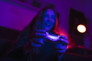 Güler yüzlü bir kızın ellerinin kumandayı tutarken ve video oyunları oynarken yakın plan görüntüsü. Geceleri renkli ışıklarla dolu bir oturma odasında..