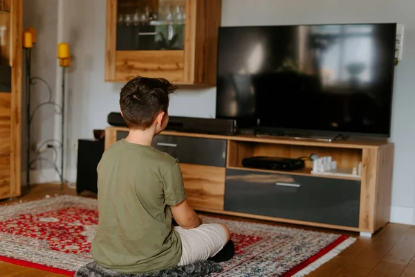 Sıradan giyinmiş, ayakları çapraz oturan ve televizyon izleyen tanınmamış bir çocuğun arka planı.