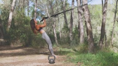 Spor elbiseli genç bir kadın spor yaparken doğadaki ağaçta sabitlenmiş spor lastiğiyle egzersiz yapıyor.