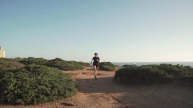 Gün batımında dağlarda spor kıyafetleri içinde koşan genç bir bayan koşucu. Deniz manzaralı. 
