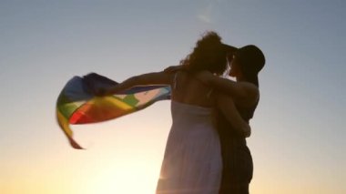 Şapkalı sıska bir bayan ve güneşli elbiseli neşeli bir kız arkadaş tatil boyunca dalgalı okyanusun kıyısında durup birbirlerine bakarken renkli gurur bayrağı çekiyorlar.