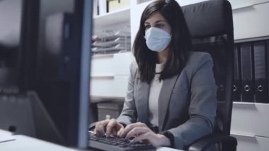 Resmi kıyafet ve koruyucu maske konusunda kendine güvenen genç bayan uzman modern çalışma alanında çalışırken masada oturuyor ve bilgisayar kullanıyor.