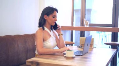 Kadın girişimci cep telefonuyla iş projesini tartışırken netbook kullanıyor ve kafedeki masada uzaktan çalışıyor.