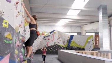 Spor kıyafetli kadın dağcı, spor salonunda antrenman yaparken yerde rehberlik ederken parlak saplı kaya duvarına tırmanıyor.