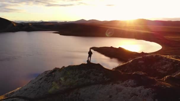 环绕着无人驾驶飞机俯瞰着冰岛日落时站在岩石上欣赏平静湖水的探险家的轮廓 — 图库视频影像
