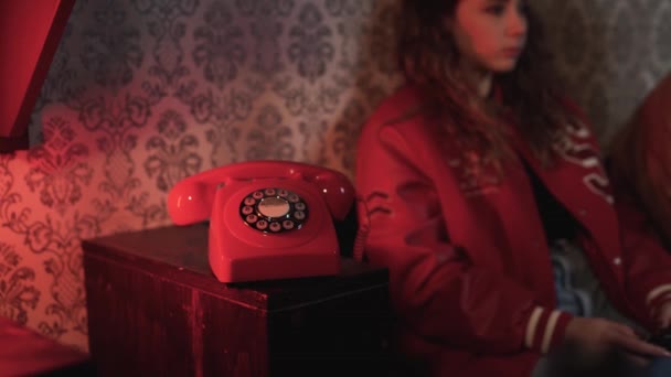 ソファーチューインガムに座ってジョイスティックを使用してビデオゲームをプレイし 古いファッションアパートで赤いレトロな電話に答える若い作物の女性のズーム — ストック動画