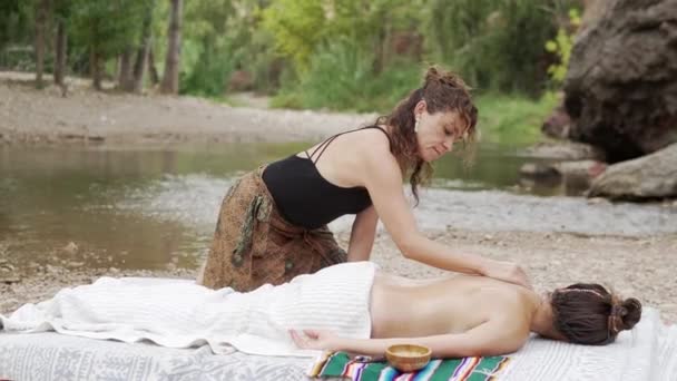 黒い上部の大人のマッサージ師の側面のビューは 池の近くの目を閉じたタオルの下にマットに横たわるトップレス女性の背中のマッサージをする — ストック動画