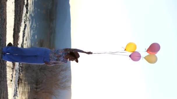 ハットでリアルタイム楽観的な女性は 日没中に湖をリッピングする近くの海岸に立っている間 カラフルな風船で踊ります — ストック動画