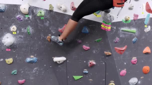 スポーツウェアやクライミングシューズで認識できない女性アスリートが ボルダリングスタジオで壁に登りながらグリップを掴む様子を下から見ると — ストック動画