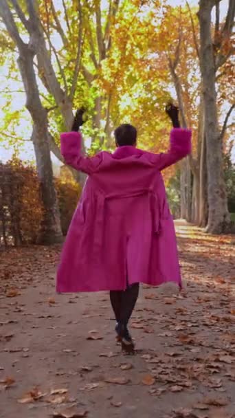 肩越しのカメラを見て頭に触れるスタイリッシュな服装のクイアモデルの追跡ショットは 秋の公園の日光の路地を歩いている間に回転します — ストック動画