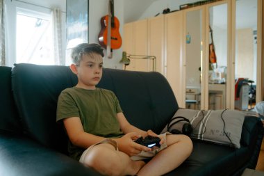 Kanepede bacak bacak üstüne atmış vaziyette oturmuş joystick ile video oyunu oynayan konsantre küçük çocuk.