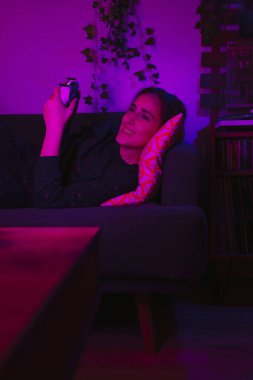 Bir kız geceleri oturma odasında pembe renkli ışıklarla video oyunu oynarken gülümsüyor..