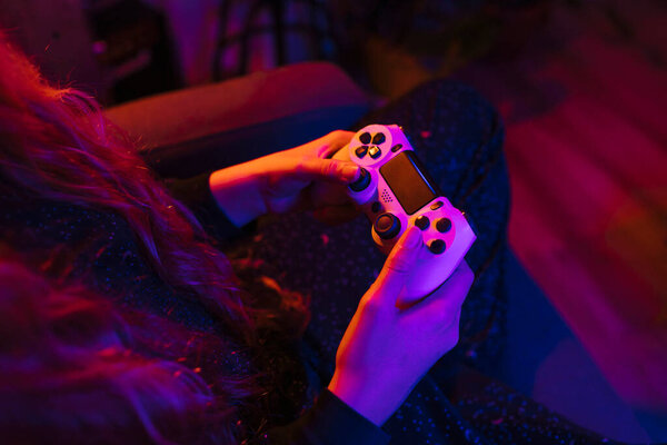 Crop from above of faceless female with long hair using gaming device while sitting with crossed legs on cozy couch in apartment in neon light