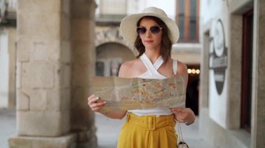 Düşünceli kadın turist şapkalı ve güneş gözlüklü. Yaz tatilinde sokakta yürürken kağıt harita okuyor.