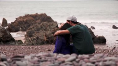 Kayalık kayalıklarla kumsalda oturan ve güneşli bir günde İrlanda 'da vakit geçirirken dalgalı deniz suyuna bakan çift.