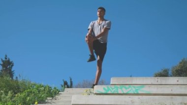 Tam boy Afrikalı Amerikalı sporcu beton merdivenlerin tepesinde duruyor ve açık havada hava egzersizi yaparken bulutsuz gökyüzüne karşı ısınma egzersizleri yapıyor.