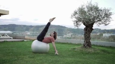 Aktif giyimli esnek bir erkek toplara yaslanıyor ve pilates eğitimi sırasında yeşil çimlerde Camatkarasana 'da yoga yapıyor.