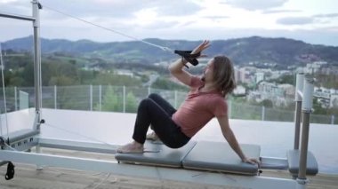 Urdhva Dhanurasana 'da pilates makinesinde kayışları olan ve terasta yoga yaparken vücudunu esneten esnek erkeğin yan görüntüsü