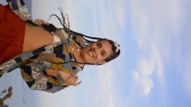 Düşük açılı, el kameralı, şık giyinen, uzun örgülü saçlarını mavi gökyüzüne doğru sallayan ve doğada dans eden enerjik, yetişkin bir kadının fotoğrafı.