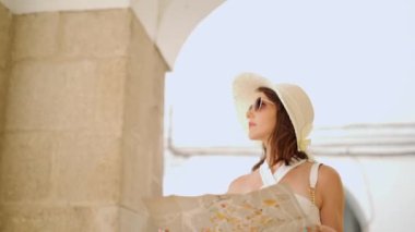Düşünceli kadın turist şapkalı ve güneş gözlüklü. Yaz tatilinde sokakta yürürken kağıt harita okuyor.