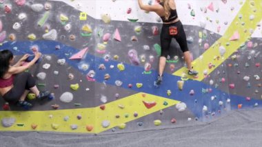 Aşağıdaki isimsiz kadınlardan, ayakkabılarına tutunarak, modern spor salonunda spor yaparken renkli sapları olan duvarlara tırmanıyorlar.