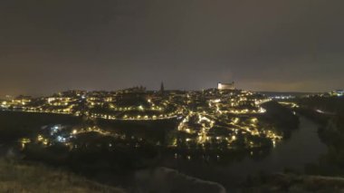Aydınlatılmış eski binaların ve kavisli yolların akşam gökyüzüne karşı tepede yer alan ve İspanya 'nın Toledo kentinde sağanak yağışı altında kalan arabaların hızlandırılmış görüntüsü