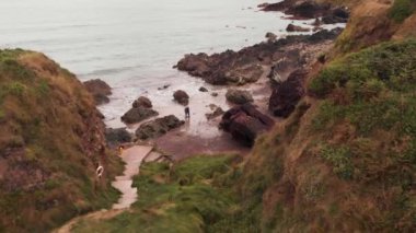 Kayalık kayalıklarla kumsalda duran, güneşli bir günde dalgalı deniz suyuna bakan ve İrlanda 'da vakit geçiren isimsiz insanların hava görüntüsü.