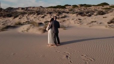 Yeni evli çiftin çıplak ayaklarıyla, gün batımında deniz kenarında kumulların arasında dans ederken nazikçe kucaklaşıp, sarmaş dolaş.