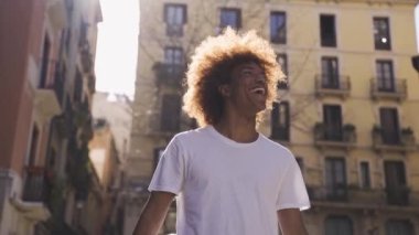 El kamerasıyla çekilmiş Afro saçlı, beyaz tişörtlü, Afro saçlı ve binaların yakınında yürürken yüzünü çeviren neşeli Afro-Amerikan erkek fotoğrafından.