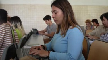 Masada oturan ve üniversitedeki bir sınıfta dizüstü bilgisayar kullanarak proje üzerinde çalışan günlük kıyafetli genç bir kadının yan görüntüsü.