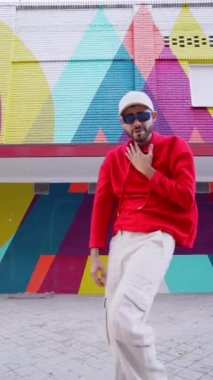 Şehir caddesindeki renkli binaya karşı dans eden şık kırmızı kapüşonlu bir erkekten.